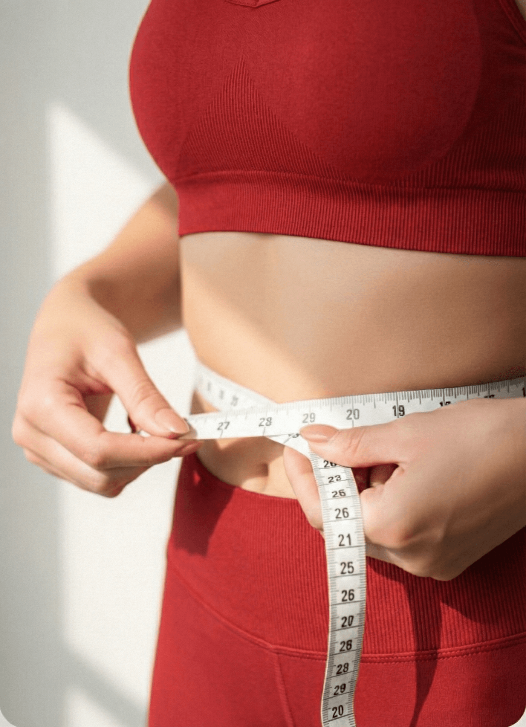 A close-up shot of a person in light blue athletic wear measuring their waist with a white measuring tape shows 28 inches, set against a bright, minimalist background with soft sunlight.
